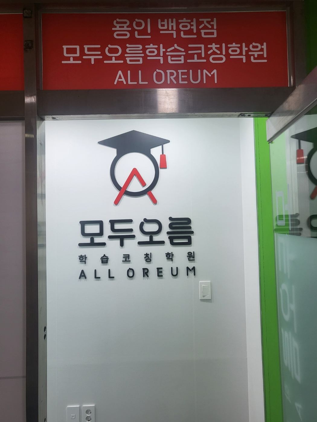 용인백현점 입구 — 흰 벽에 학사모를 쓴 원형 로고와 모두오름 학습코칭학원 ALL OREUM 글자가 붙어 있고, 위쪽 빨간 간판에 용인 백현점 모두오름학습코칭학원이라고 적혀 있다