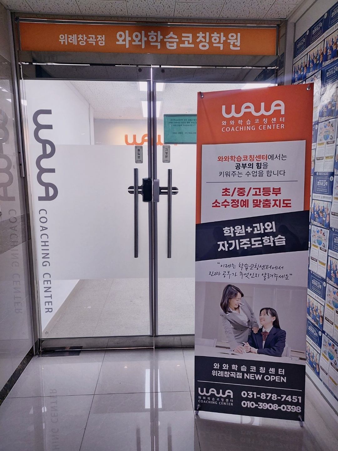 위례창곡점 출입구 — 유리문 위에 '위례창곡점 와와학습코칭학원'이라고 적힌 주황색 간판이 있고, 유리문에는 WAWA COACHING CENTER 로고가 붙어 있다. 문 오른쪽에는 '학원+과외 자기주도학습', '초/중/고등부 소수정예 맞춤지도'라고 적힌 주황색 안내 배너가 세워져 있다