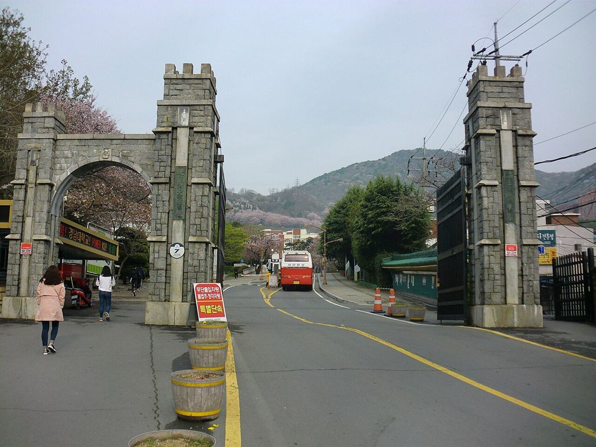 경남 창원시 마산합포구 경남대학교 정문 — 돌로 쌓은 아치형 문기둥과 그 뒤로 이어지는 캠퍼스 길, 벚꽃이 핀 산