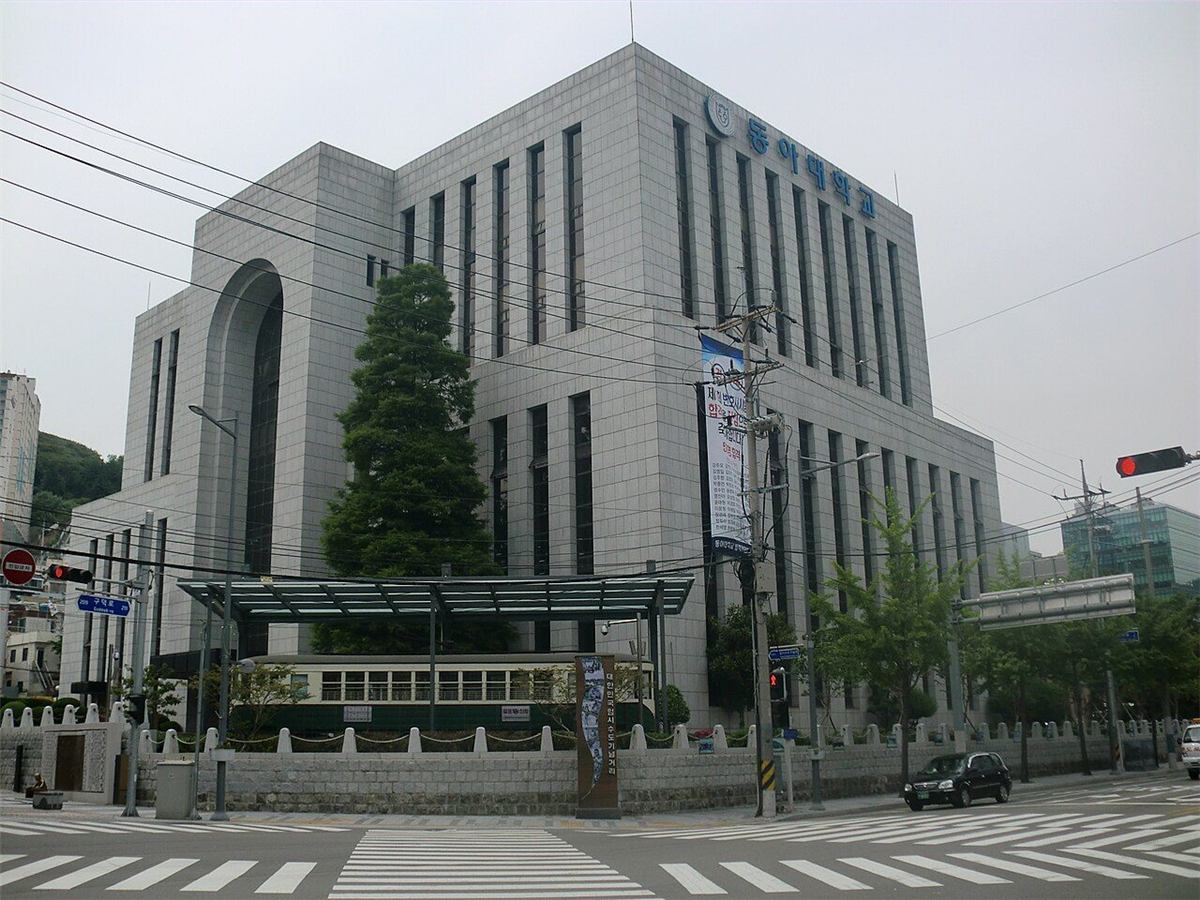 동아대학교 부민캠퍼스 — 교차로 모퉁이에 선 회색 석재 외벽의 각진 건물로, 위쪽에 동아대학교 교표와 파란 글자 간판이 붙어 있다. 건물 앞 낮은 돌담 안쪽에는 유리 지붕을 씌운 옛 전차 한 량이 전시돼 있고, 앞쪽으로 횡단보도와 신호등이 보인다
