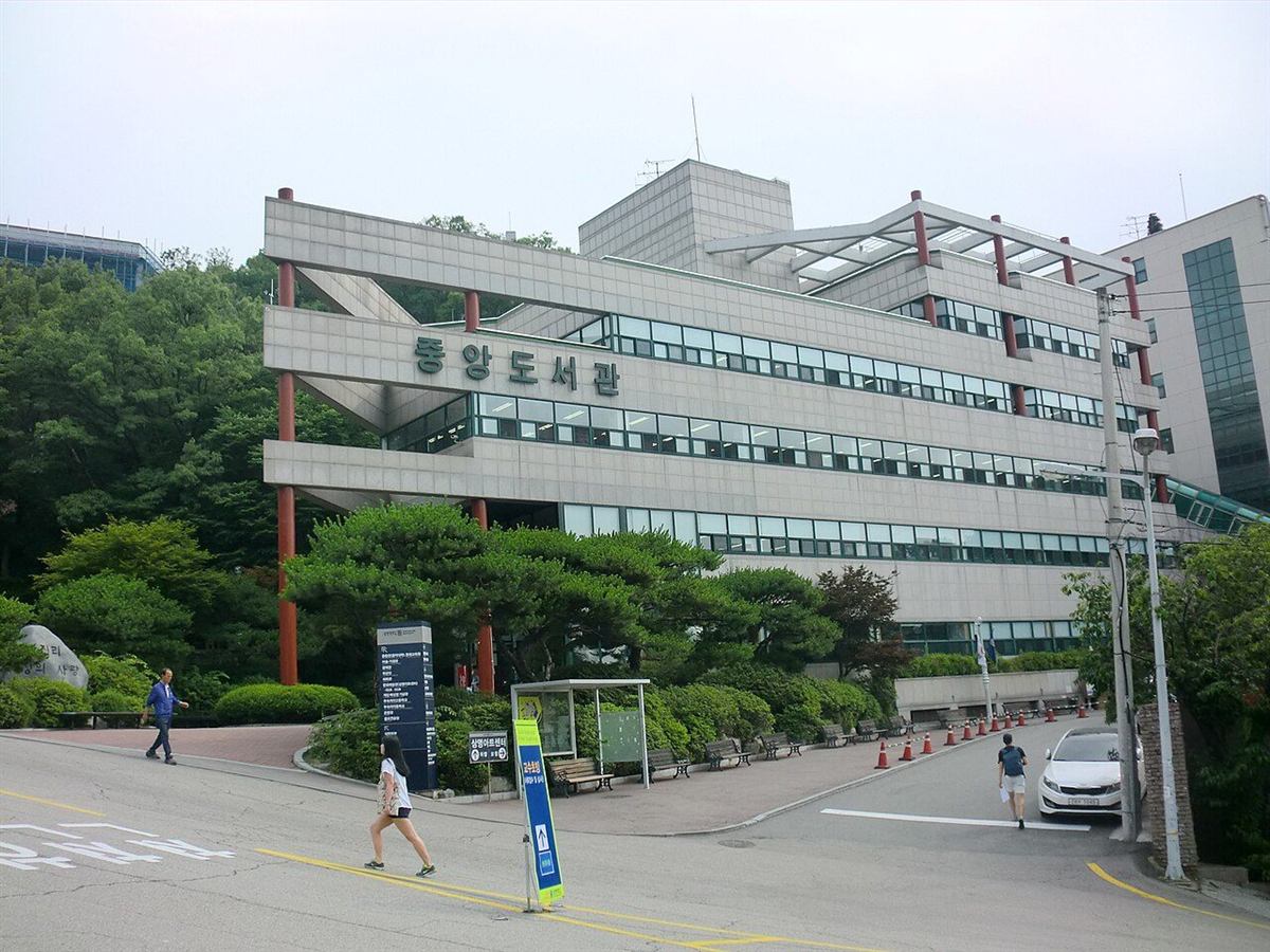 상명대학교 서울캠퍼스 중앙도서관 — 언덕을 따라 계단식으로 올라가는 회색 콘크리트 건물에 붉은 기둥이 세로로 서 있고, 건물 정면 위쪽에 중앙도서관이라는 큰 글자가 붙어 있다. 건물 앞으로는 소나무와 관목이 심긴 화단과 넓은 진입 도로가 보인다