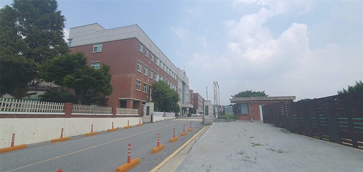 국립한국교통대학교 의왕캠퍼스 입구 — 붉은 벽돌 외벽에 회색 지붕을 얹은 긴 강의동 건물이 왼쪽으로 늘어서 있고, 그 앞으로 진입 도로와 보도블록이 이어진다. 오른쪽에는 낮은 경비실 건물과 세로로 선 안내탑이 보인다