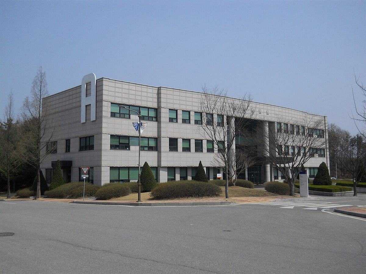 한국기술교육대학교 대학본부 — 회색 석재로 마감된 3층 건물이 도로 모퉁이에 서 있고, 왼쪽 옥상에 흰색 조형 구조물이 올라가 있다. 건물 앞으로는 다듬어진 관목 화단과 가로등, 넓은 진입 도로가 보인다